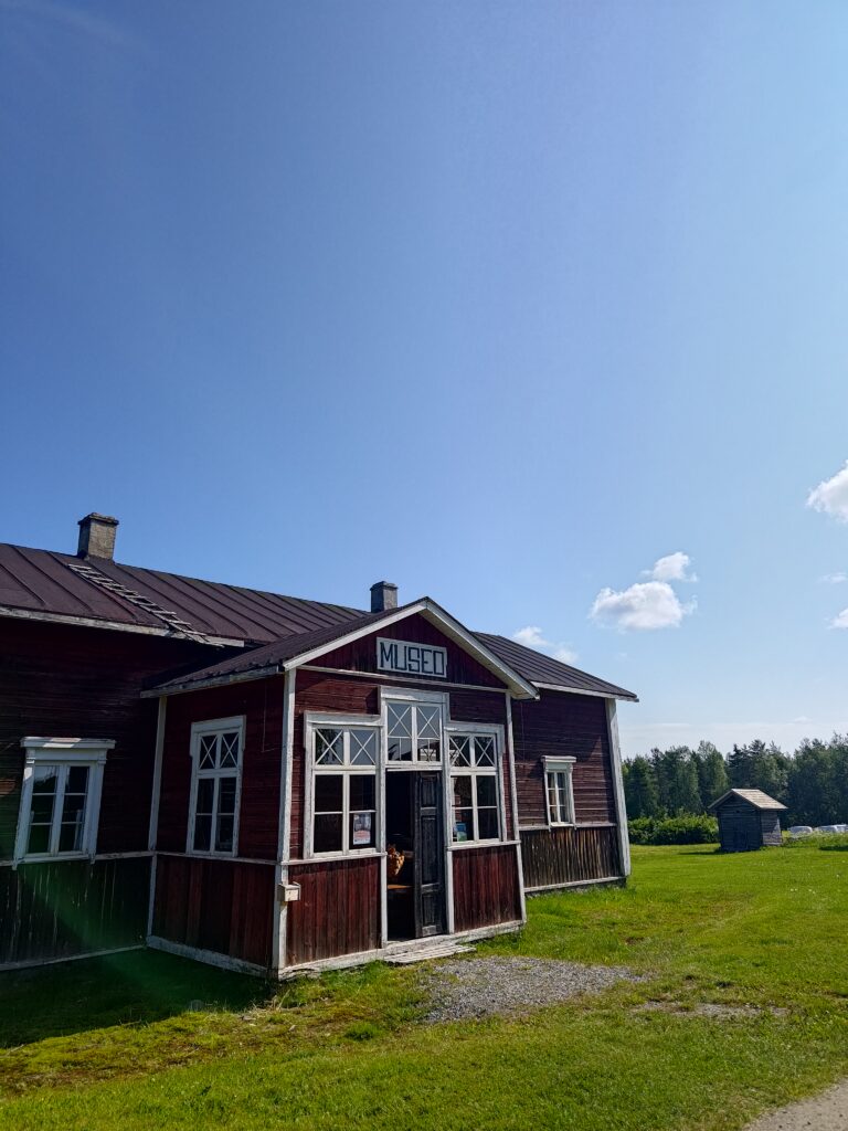 Posion kotiseutumuseo ulkoa. Rakennus on punainen hirsirakennus, oviaukon yllä itsetehty kyltti, jossa lukee "Museo". Kuvan ottohetkellä sää on ollu aurinkoinen ja miltei pilvetön. Vihreä ruoho, punainen rakennus ja sininen taivas luovat hienon kontrastin.